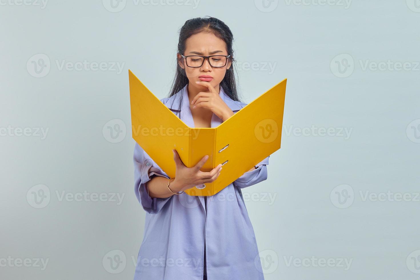 Portrait of serious young Asian business woman holding chin, contemplating something while holding document folder isolated on purple background photo