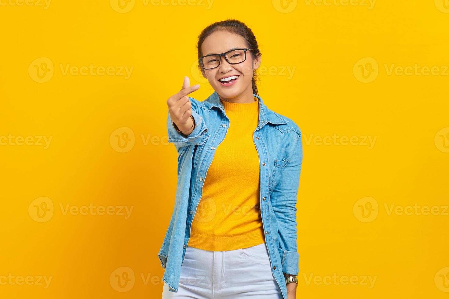 Portrait of smiling young Asian woman student in denim clothes showing korean heart with two fingers crossed isolated on yellow background. People sincere emotions lifestyle concept photo