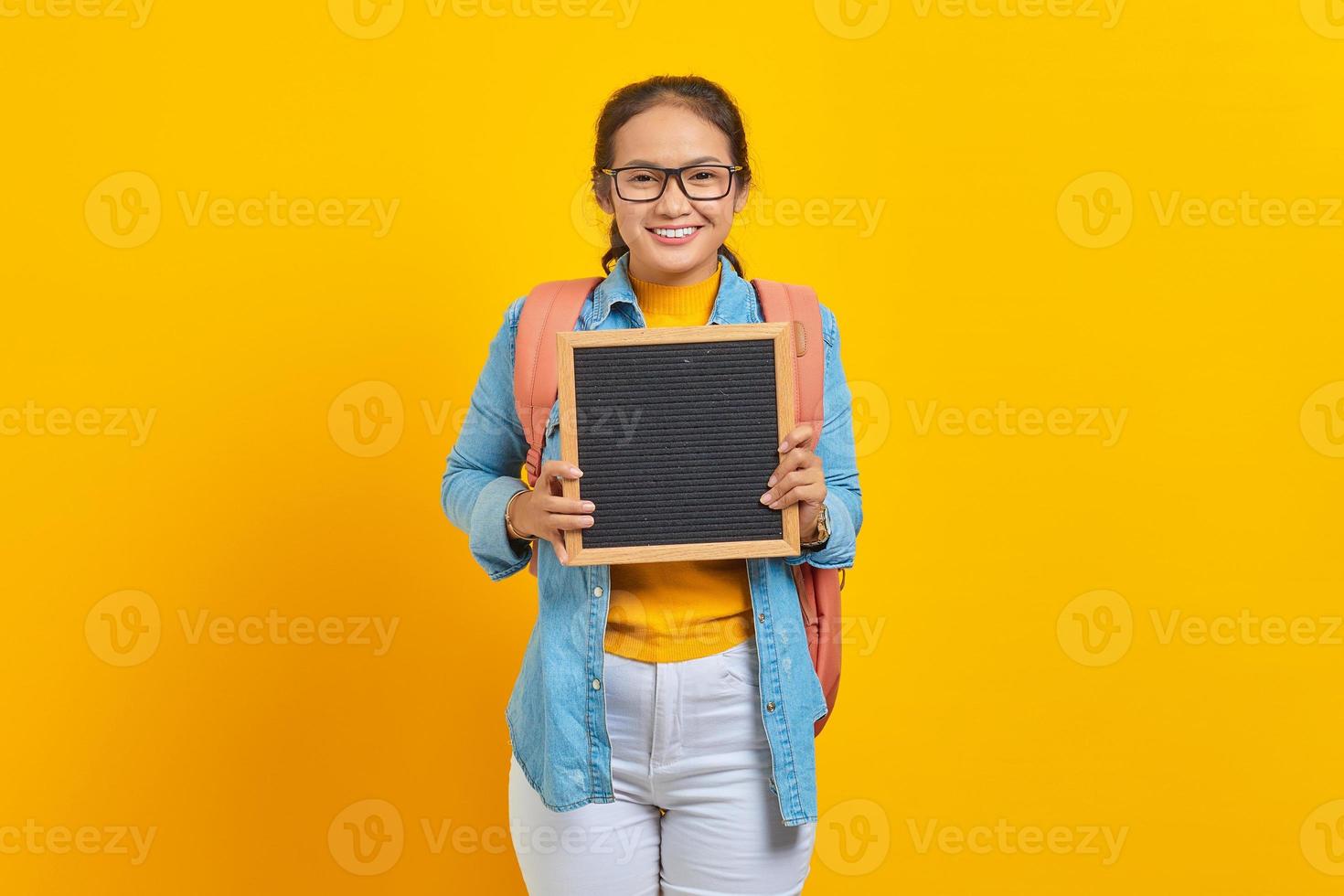 retrato de una joven estudiante asiática sonriente vestida de forma informal con mochila sosteniendo una pizarra en blanco aislada de fondo amarillo. educación en concepto de universidad universitaria foto