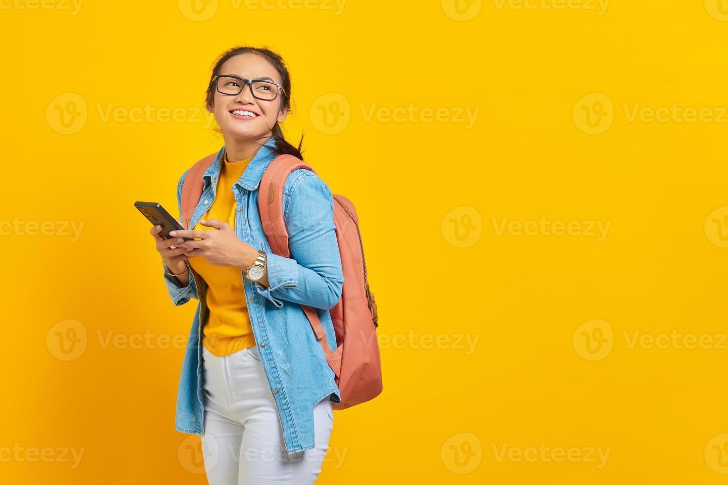 Portrait of smiling young Asian woman student in casual clothes with backpack using mobile phone and looking aside isolated on yellow background. Education in college university concept photo