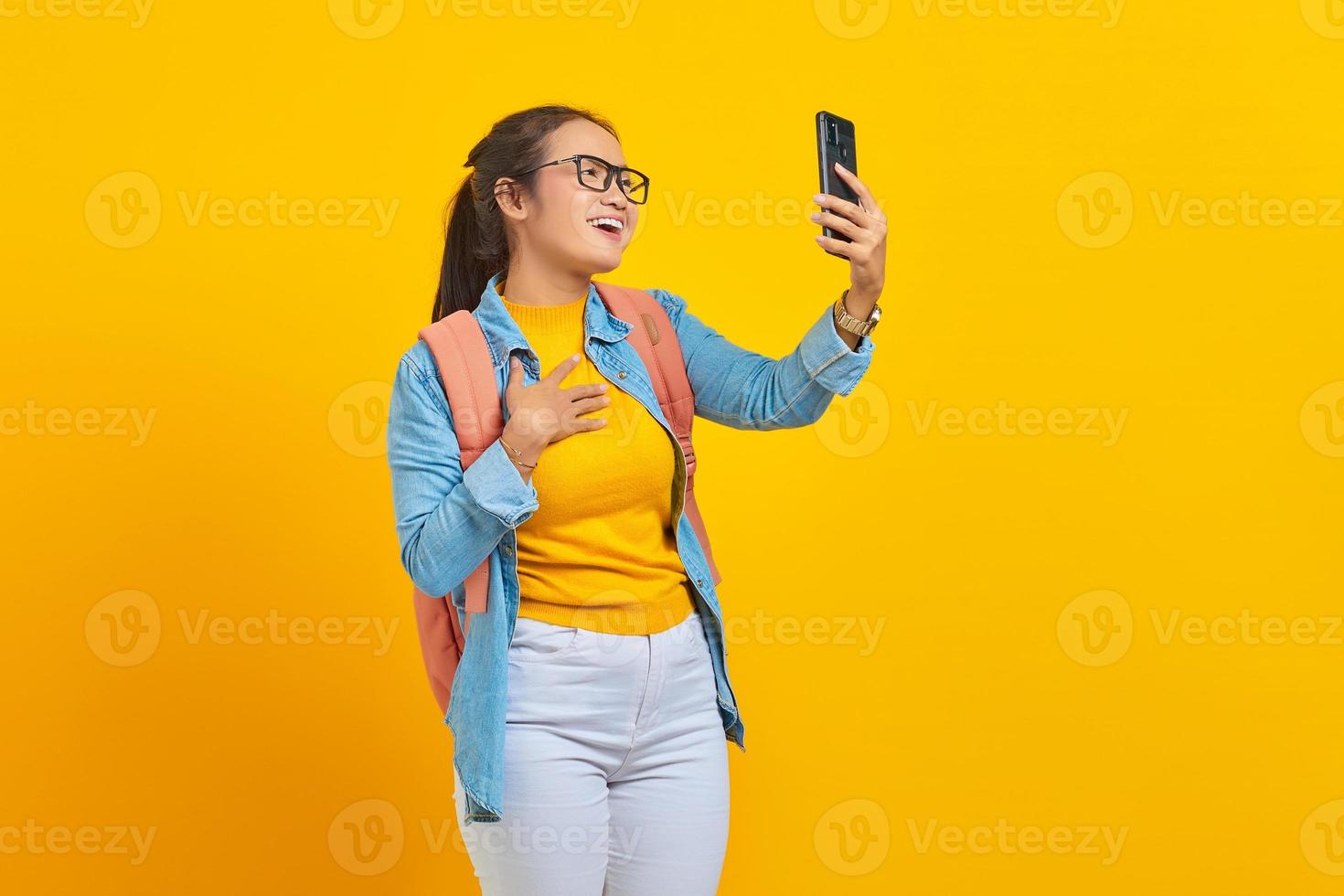 Portrait of  cheerful young Asian woman student in casual clothes with backpack having video call with smartphone and hands on chest isolated on yellow background photo
