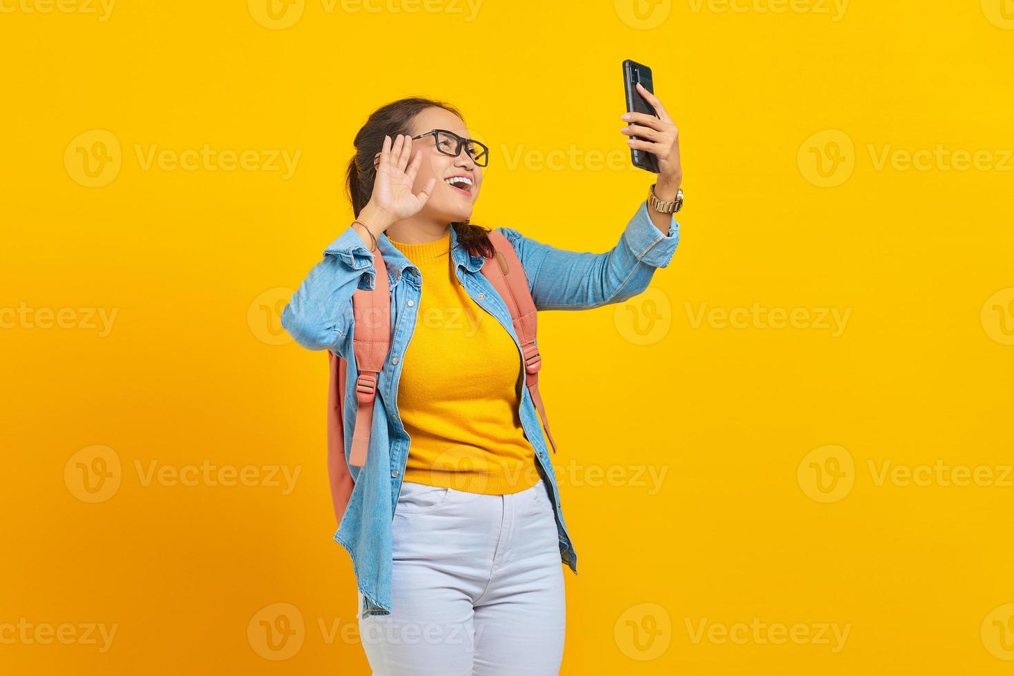 Portrait of  cheerful young Asian woman student in casual clothes with backpack having video call and waving a hand at smartphone camera isolated on yellow background photo