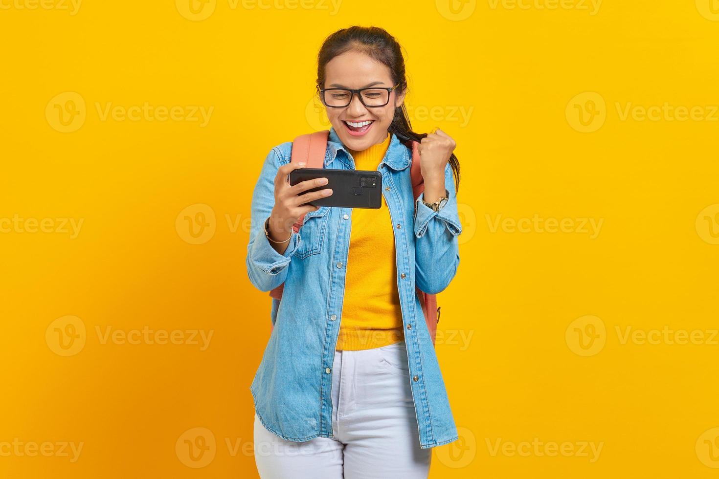 retrato de una alegre joven estudiante asiática vestida de forma informal con mochila jugando videojuegos en el teléfono móvil y celebrando el éxito aislada de fondo amarillo foto