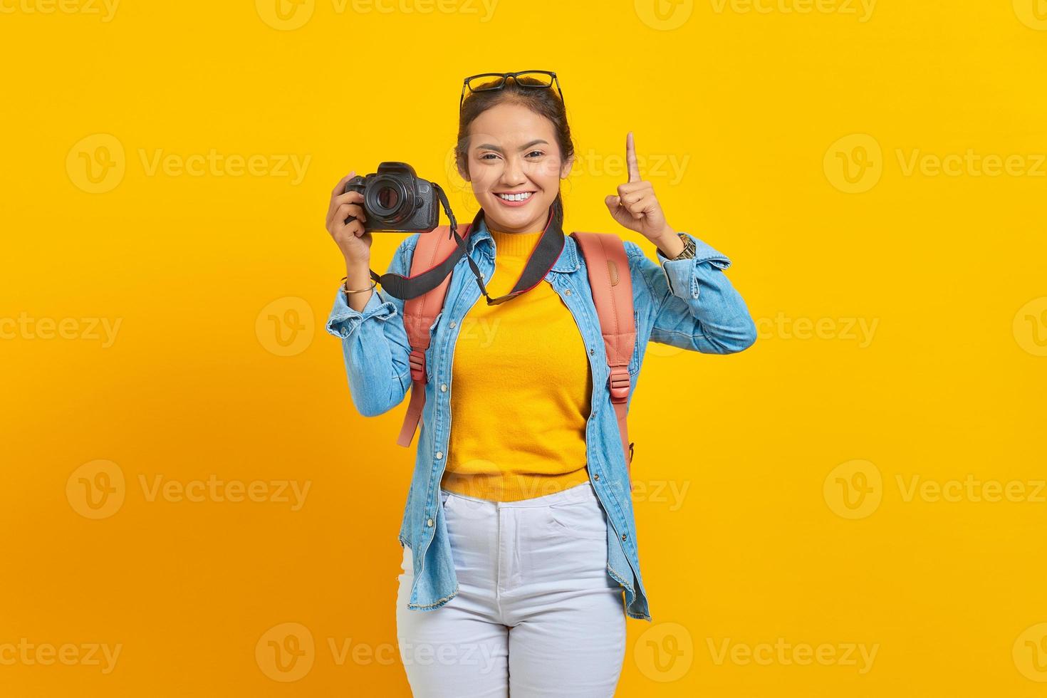Portrait of smiling young Asian woman in denim clothes with backpack holding professional camera and pointing up with finger isolated on yellow background photo