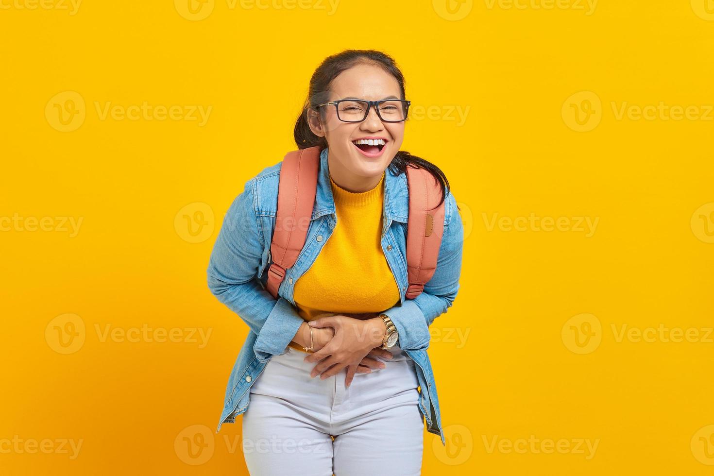 Beautiful young Asian woman student in denim outfit with backpack showing loud laughter with hand on stomach isolated on yellow background. Education in university concept photo