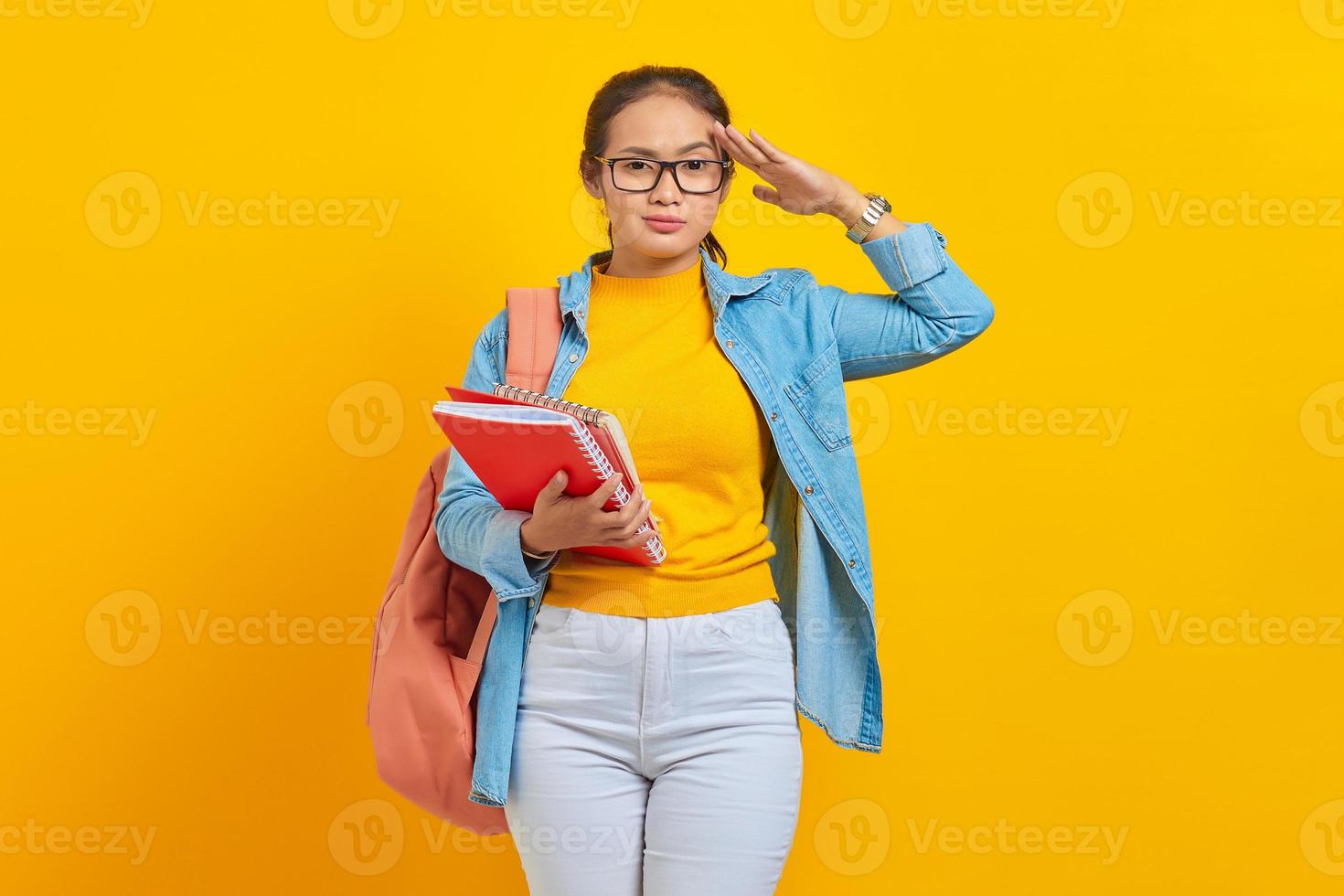 hermosa joven estudiante vestida de denim con mochila, sosteniendo un cuaderno y mostrando un gesto de respeto con la mano aislada en el fondo amarillo. educación en el concepto de colegio universitario de secundaria foto