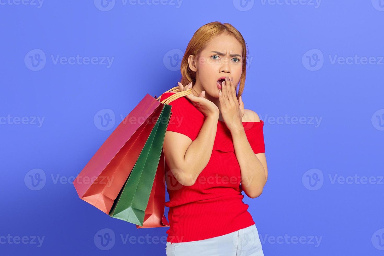 Surprised young Asian woman holding shopping bags with open mouth and looking at camera isolated over purple background photo