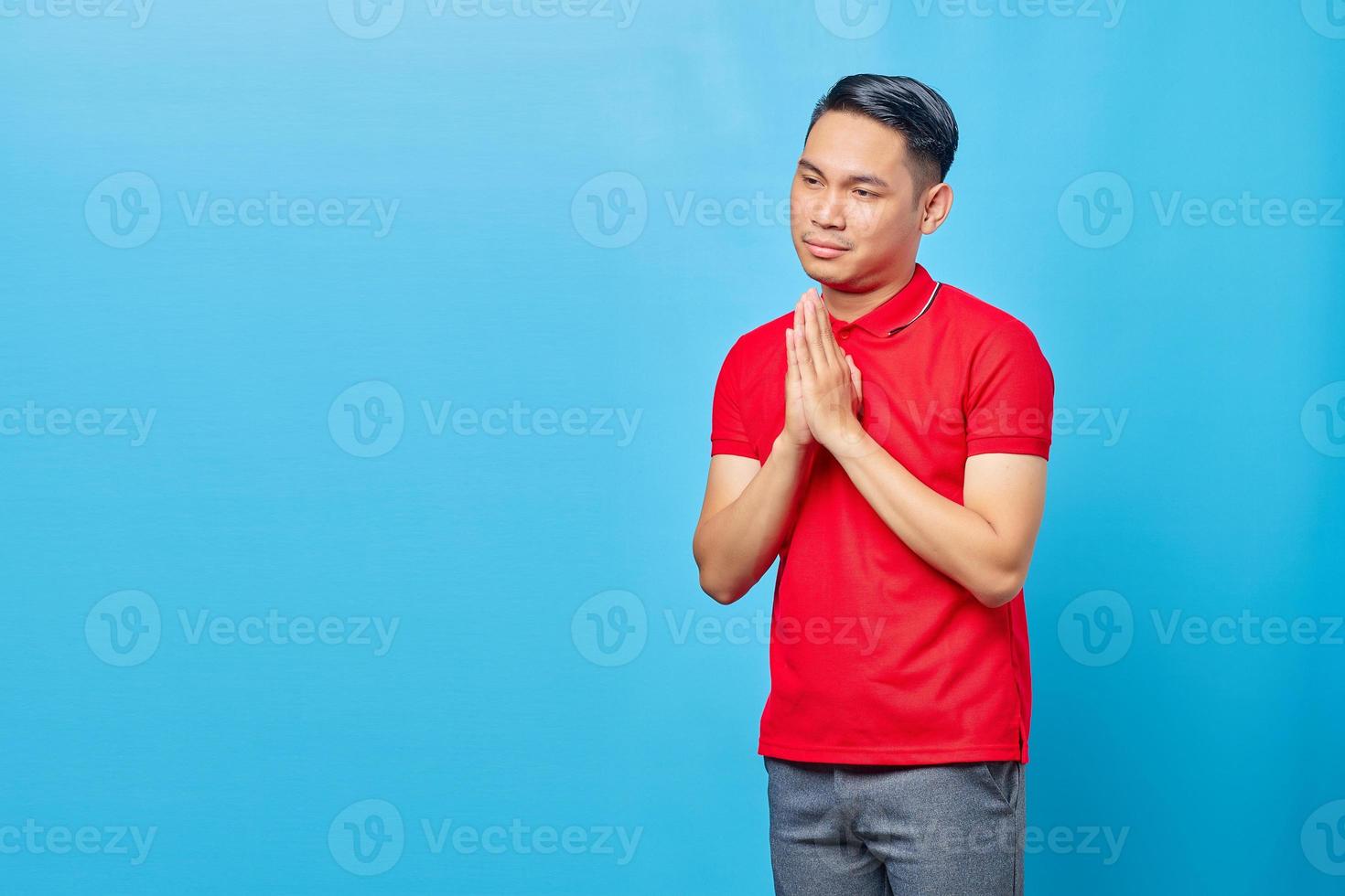 Portrait of handsome Asian young man wearing red shirt praying with hands together asking for forgiveness isolated on blue background photo