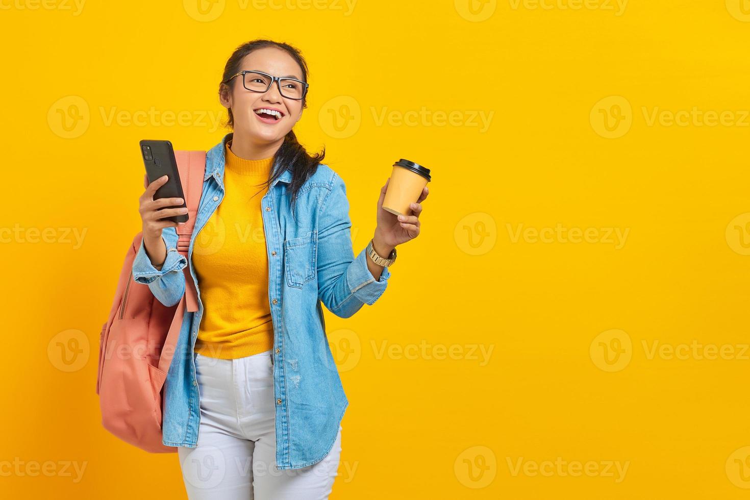 retrato de una alegre joven estudiante asiática vestida de denim con mochila sosteniendo una taza de café y un smartphone aislado de fondo amarillo foto