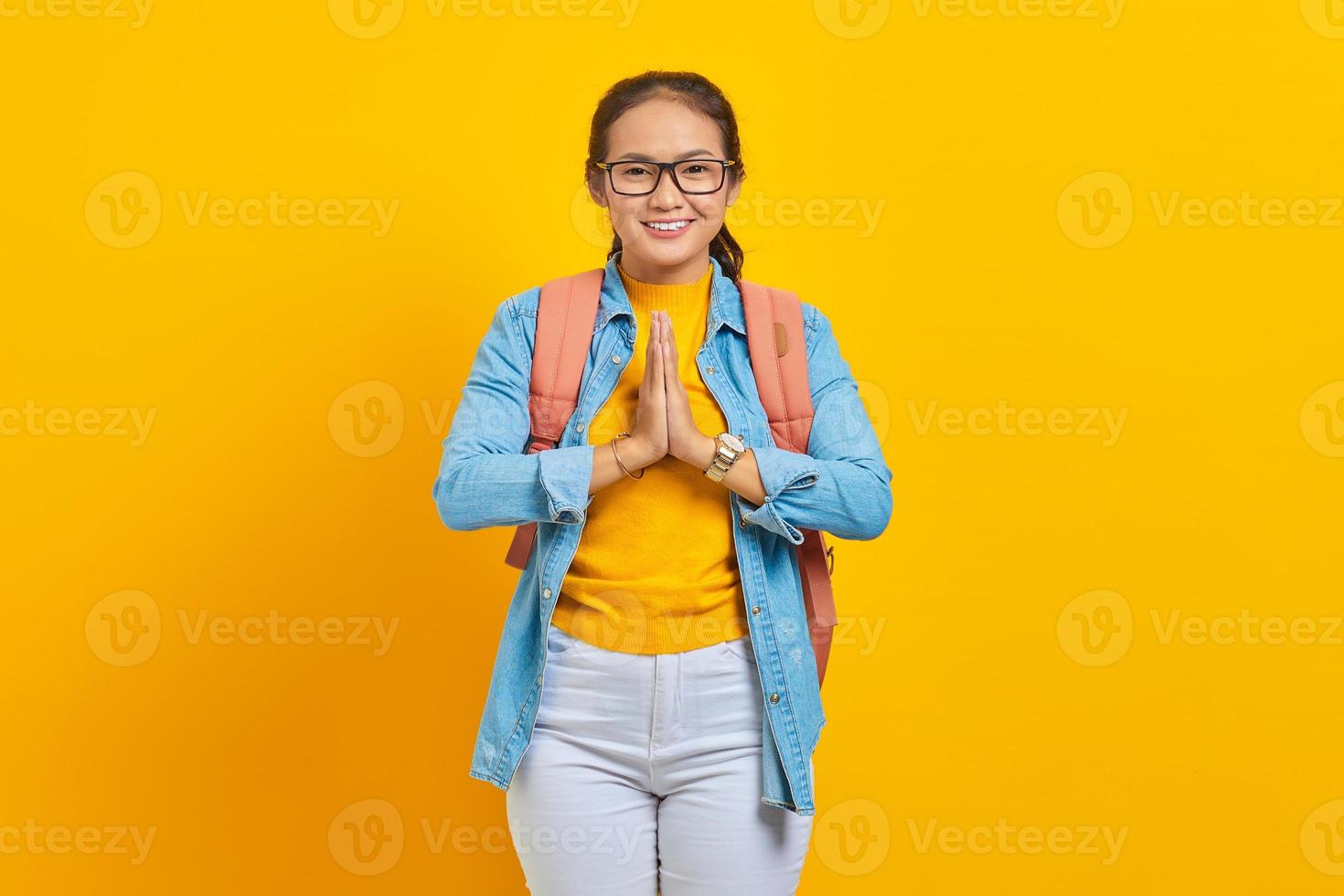 Smiling young Asian woman student in denim outfit with backpack looking confident, open sign to greet and greet customers isolated on yellow background.  Education in university college concept photo