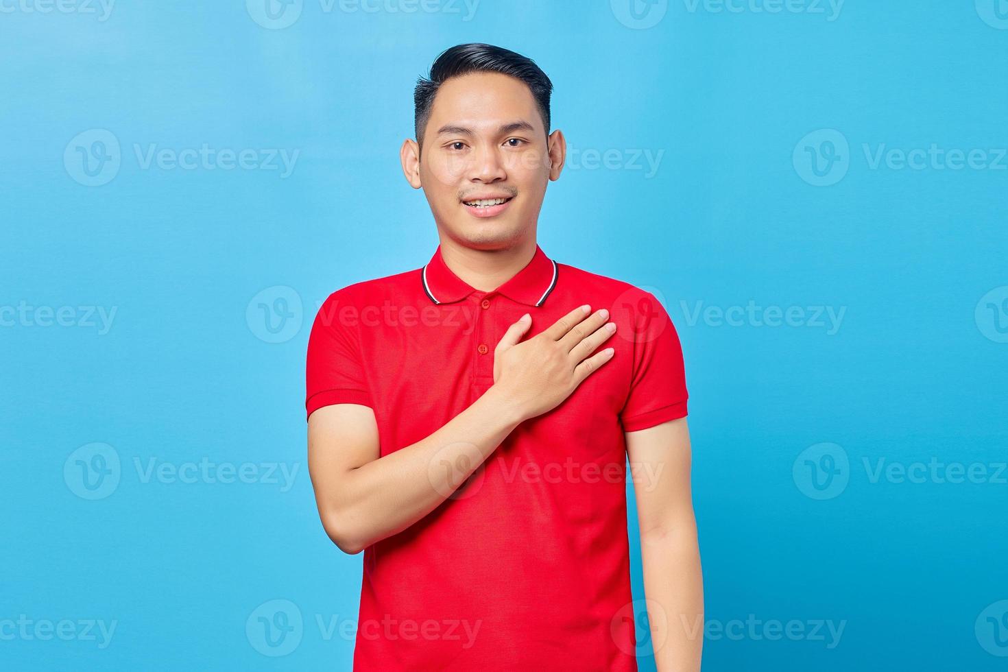 Portrait of handsome asian young man in red shirt holding hands on chest to thank someone and express gratitude isolated on blue background photo