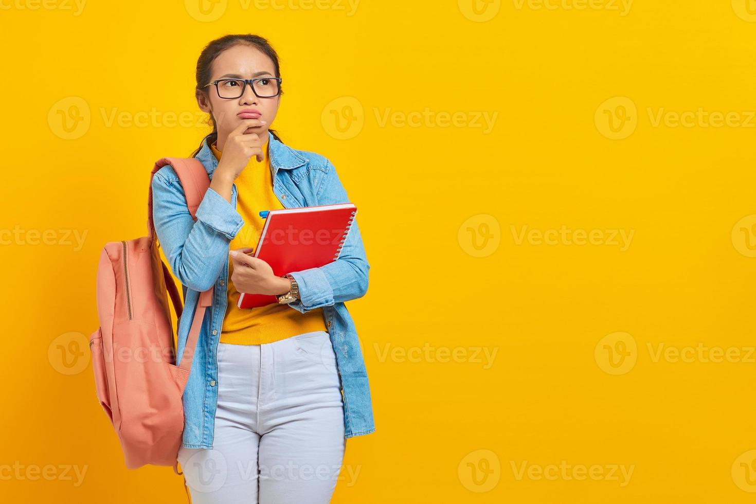 retrato de una joven estudiante asiática pensativa vestida de forma informal con mochila sosteniendo un libro y pensando en una pregunta aislada en un fondo amarillo. educación en concepto de universidad universitaria foto