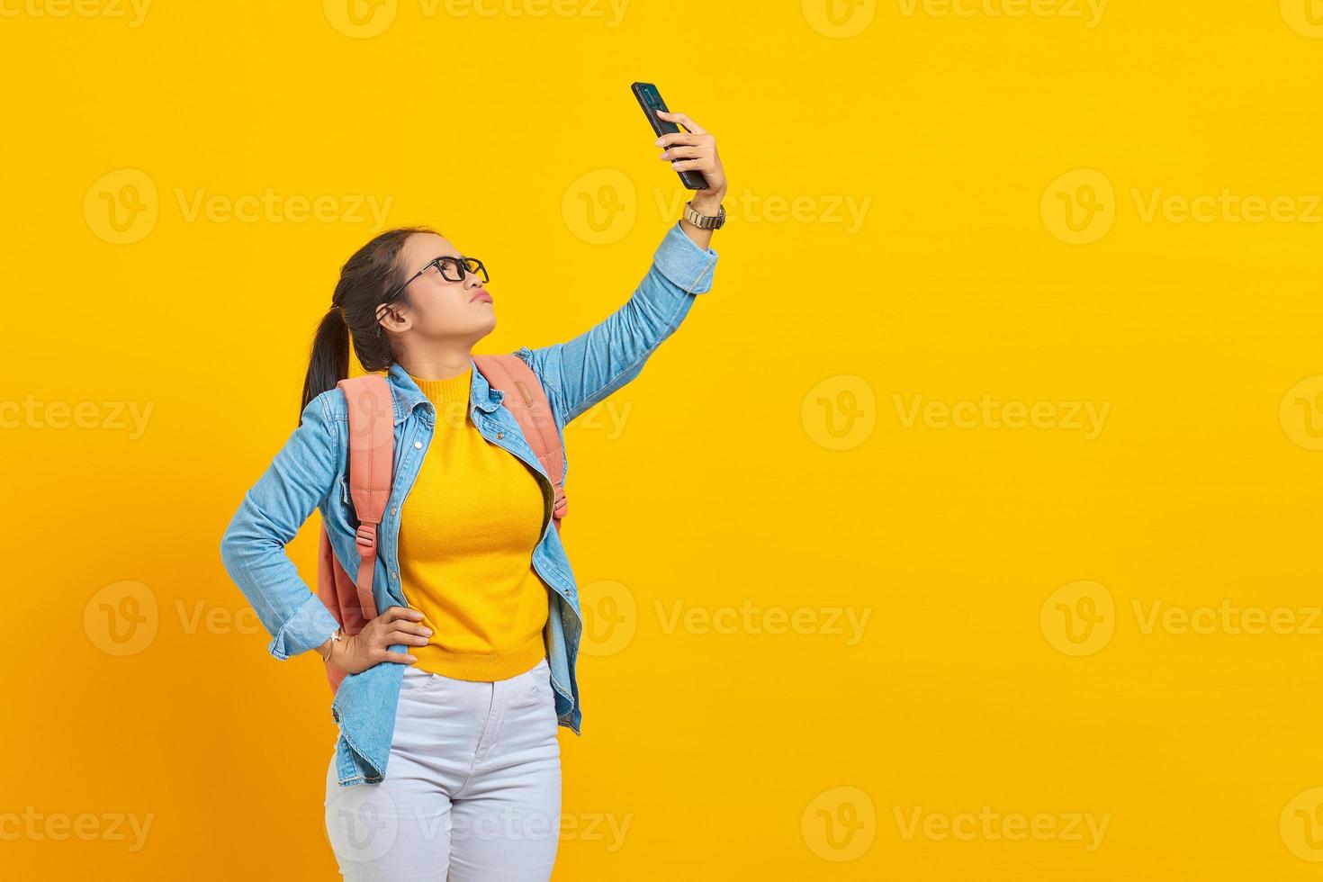 retrato de una joven estudiante asiática confundida con ropa informal con mochila usando un smartphone y buscando una red de Internet aislada en un fondo amarillo. educación en concepto de universidad universitaria foto