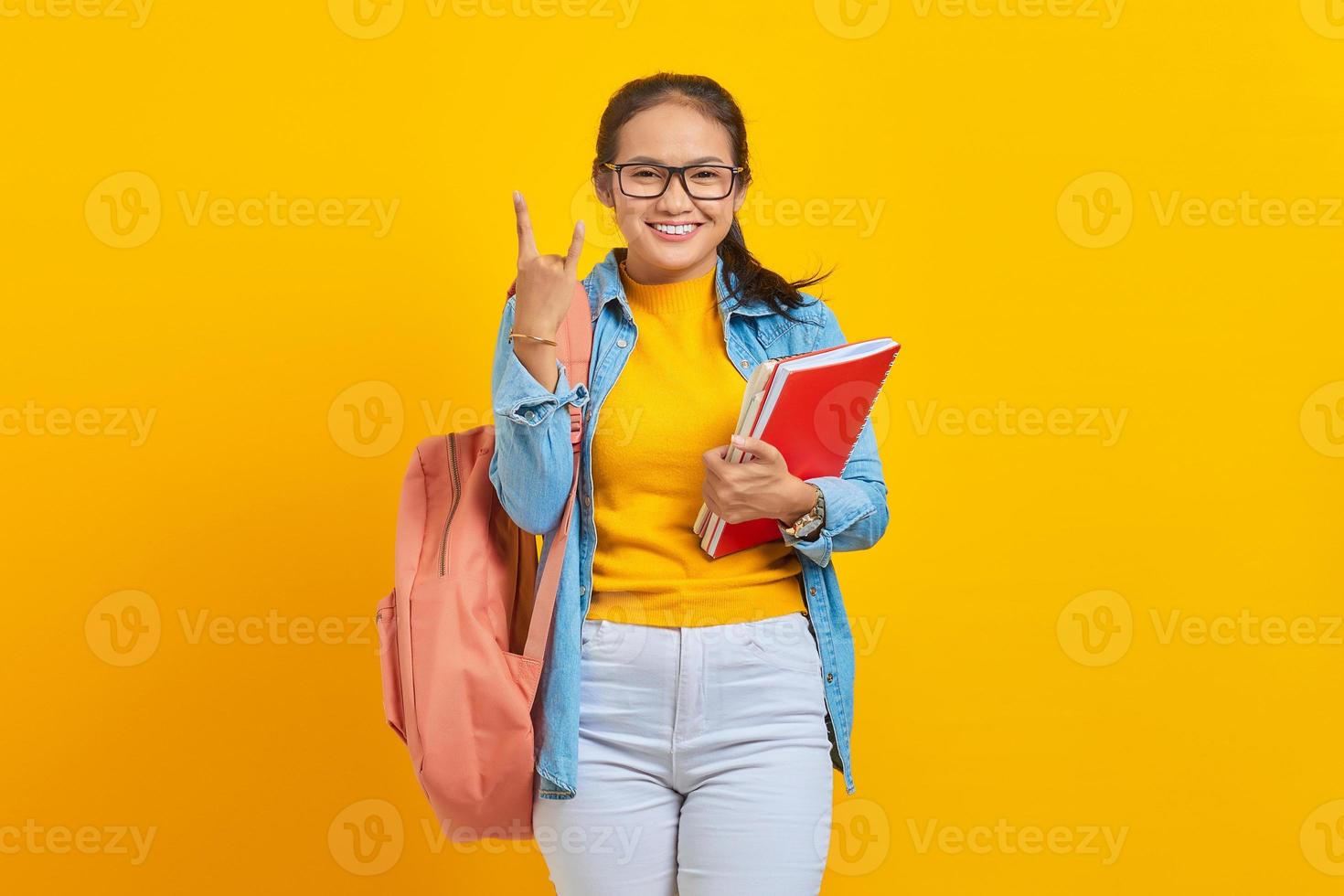 retrato de un hermoso joven estudiante asiático vestido con ropa de mezclilla con mochila sosteniendo un cuaderno y gritando con una expresión loca haciendo un símbolo de roca con las manos arriba aislado en un fondo amarillo foto