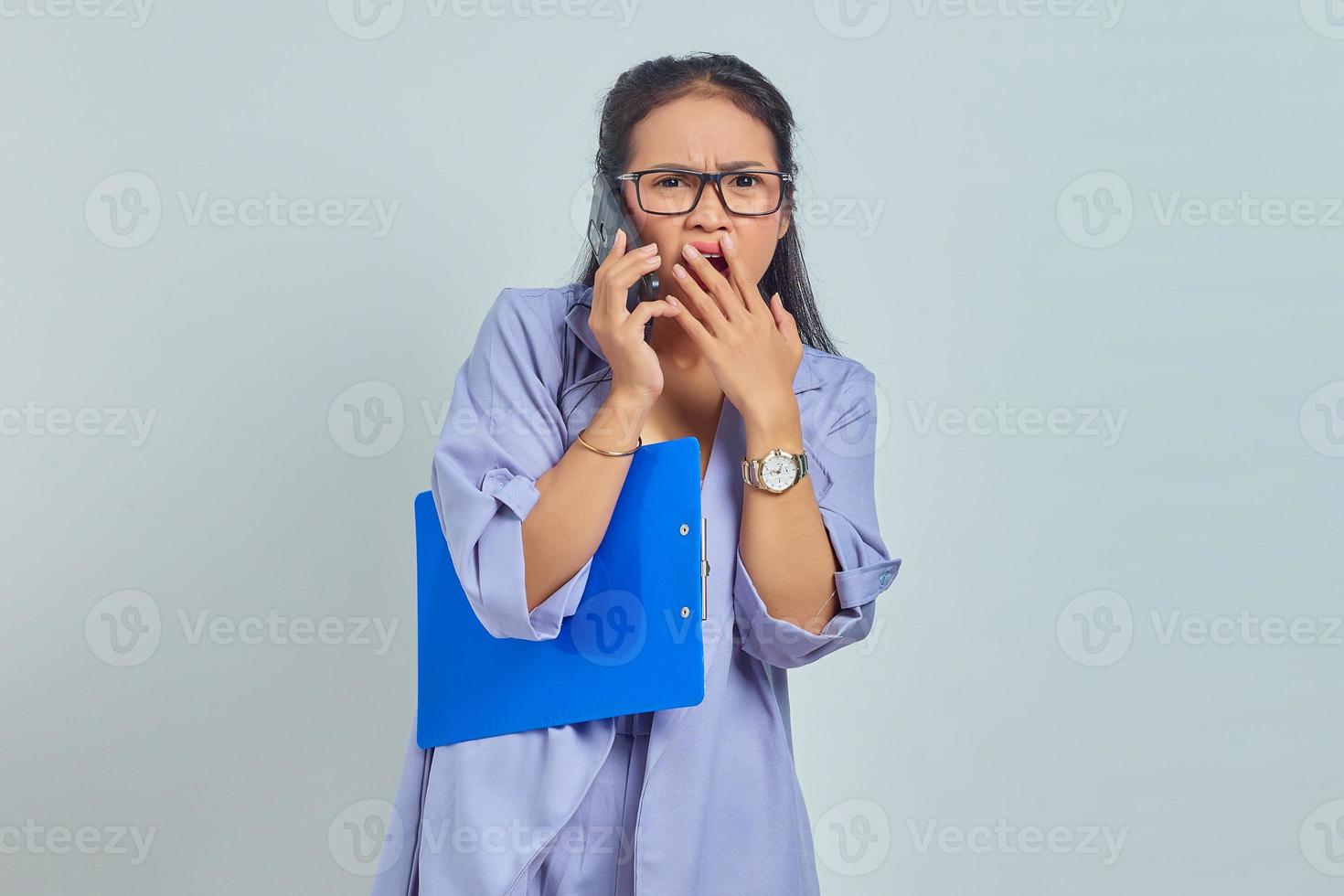 Portrait of cheerful young Asian woman laughing while talking on mobile phone with friends about something funny while holding document folder isolated on purple background photo