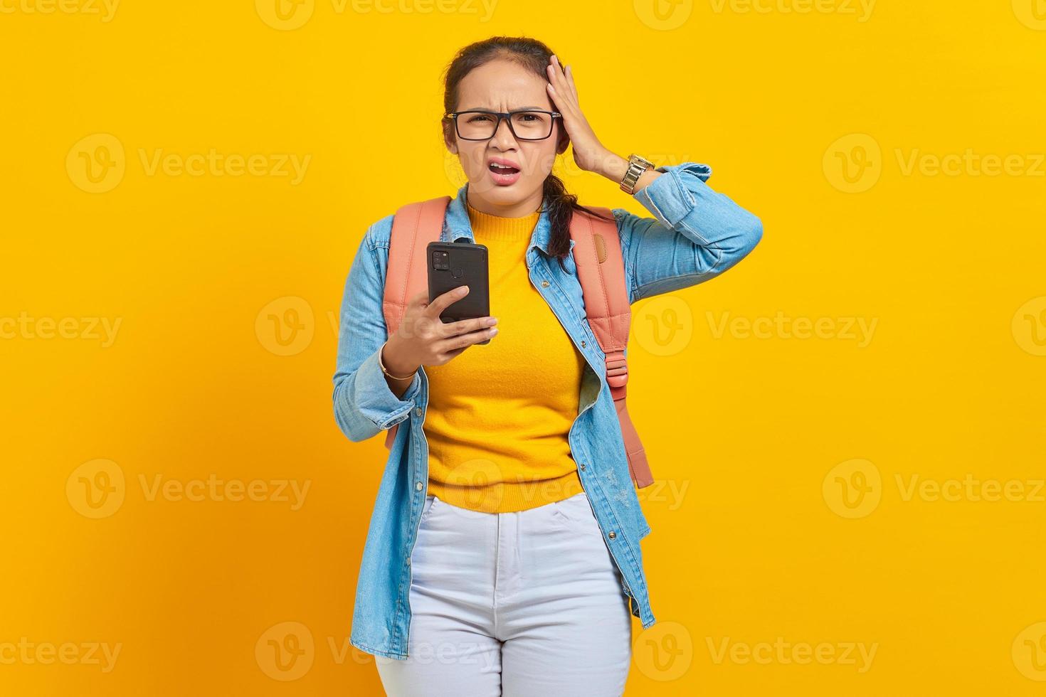 Portrait of surprised young asian woman student in casual clothes  with backpack using mobile phone with open mouth and holding hands in head isolated on yellow background photo