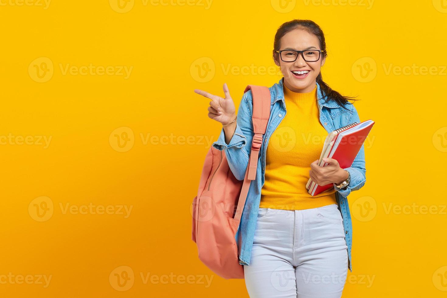 Portrait of smiling young Asian woman college student in denim clothes with backpack holding notebook and pointing finger at copy space isolated on yellow background photo