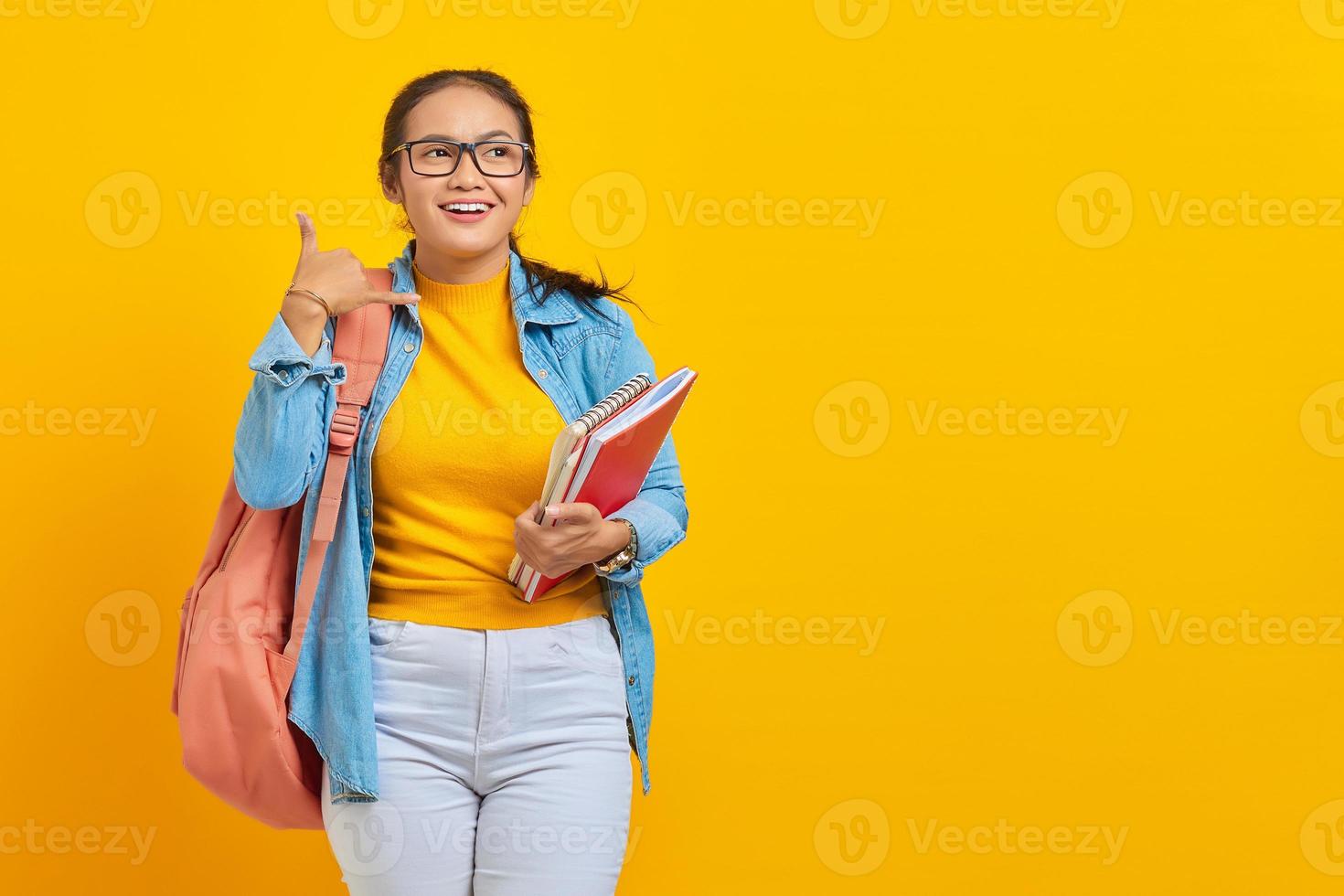 Portrait of Smiling young Asian woman student in denim clothes with backpack holding notebook and  doing phone gesture like says call me back isolated on yellow background photo