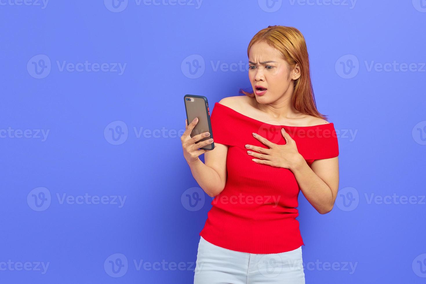 Portrait of beautiful young Asian woman in red dress using mobile phone with surprised and shocked expression isolated on purple background photo