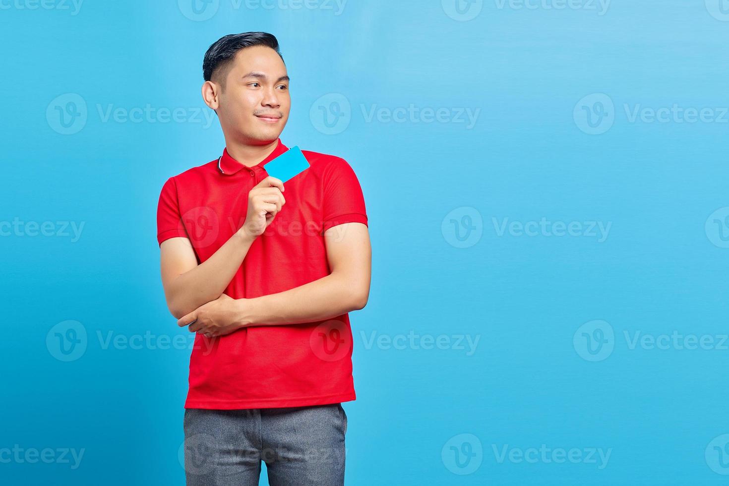 Portrait of excited Asian young man showing blank credit card and looking at copy space isolated on blue background photo