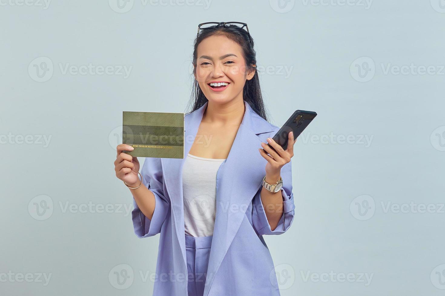 retrato de una joven asiática alegre que muestra el libro del vehículo y usa un teléfono móvil aislado de fondo blanco foto