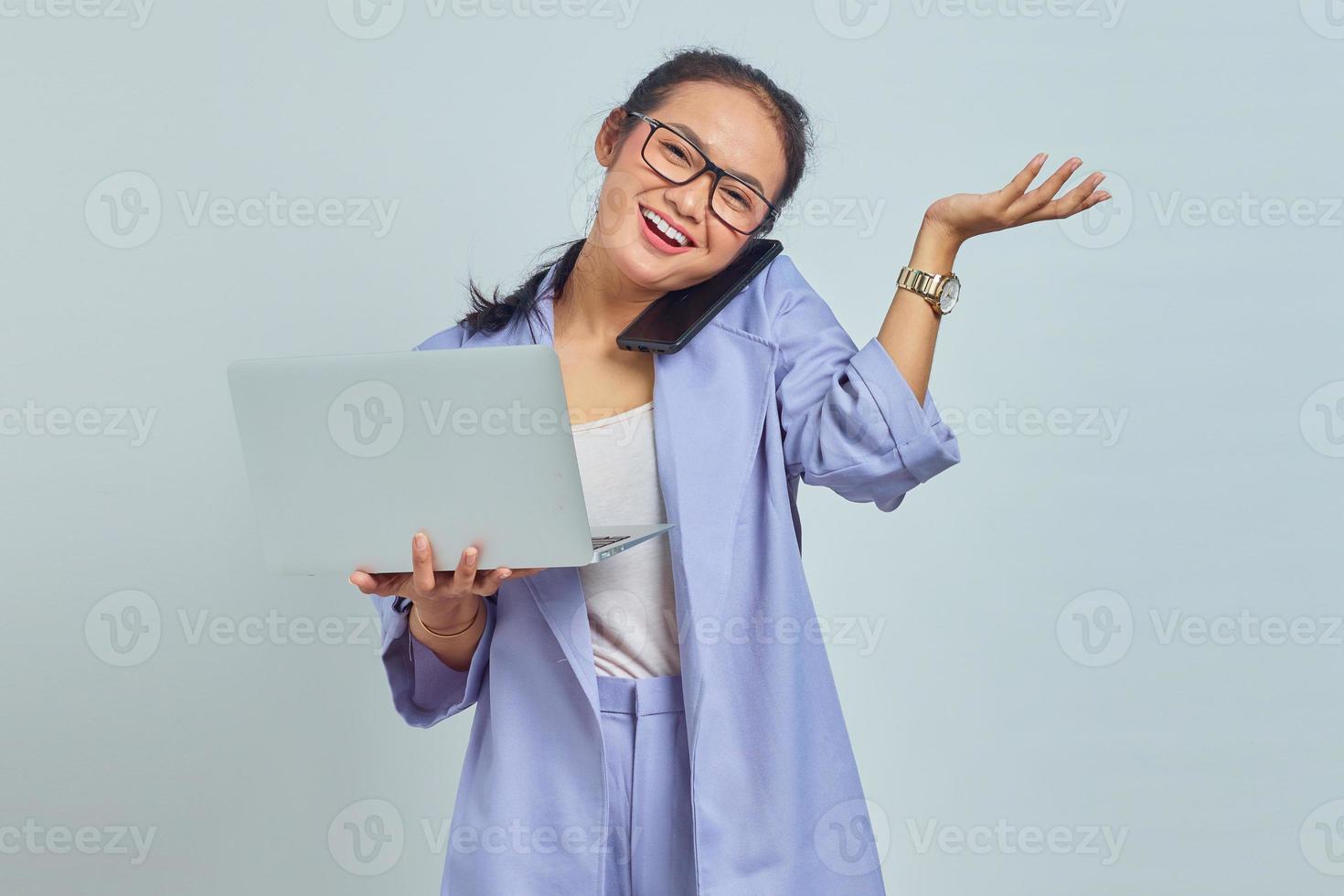 Portrait of smiling young Asian woman talking on mobile phone and holding laptop, pointing to copy space with palms isolated on white background photo