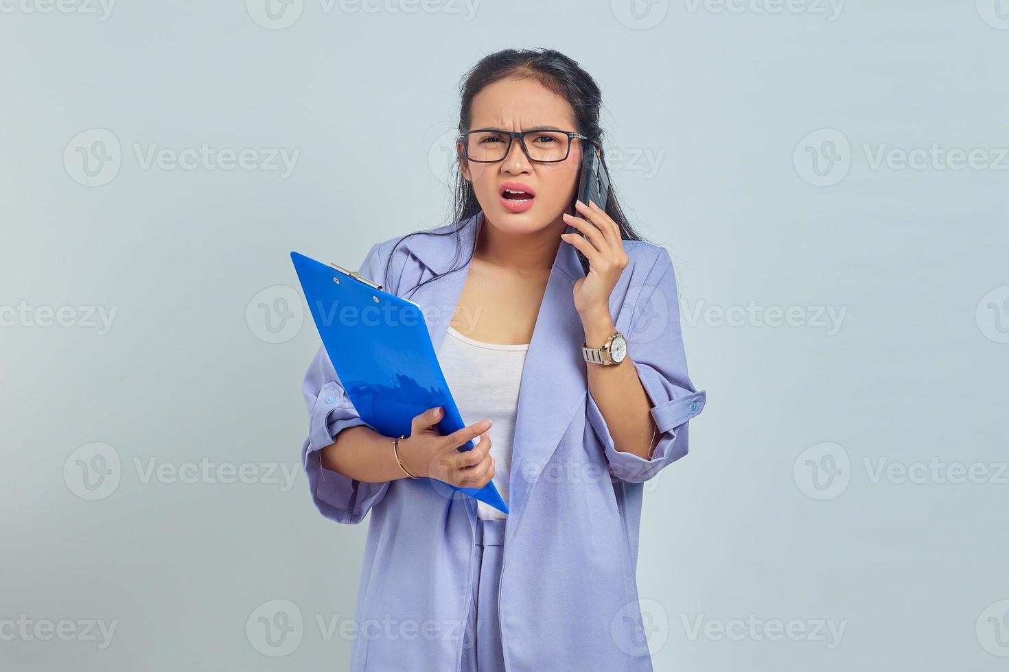 Portrait of surprised young Asian woman laughing while talking on mobile phone with friends about something funny while holding document folder isolated on purple background photo