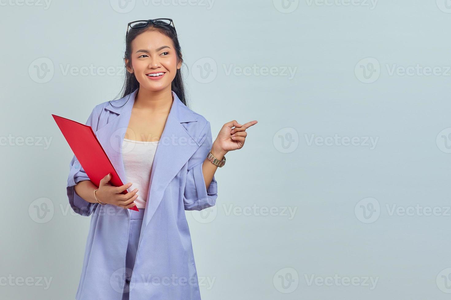 retrato de una joven mujer de negocios asiática sonriente con traje sosteniendo una carpeta de documentos y señalando con el dedo el espacio de copia aislado en un fondo morado foto