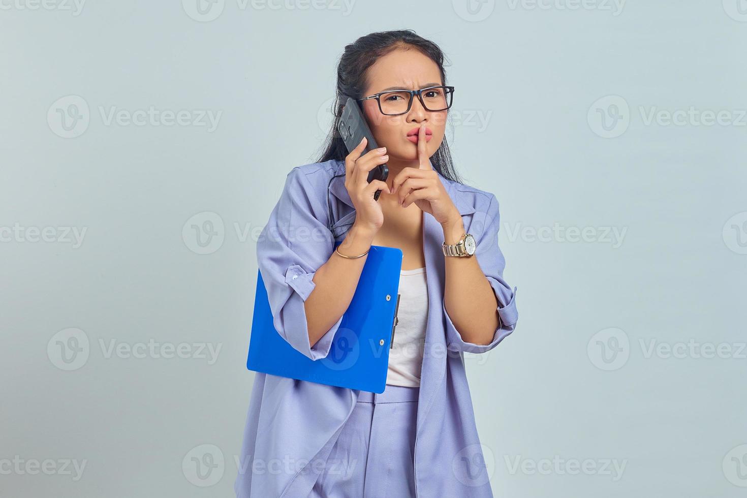 Portrait of beautiful young Asian woman talking on mobile phone and making silent gesture isolated on purple background photo