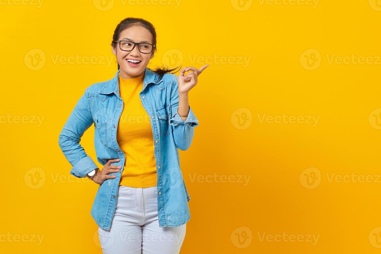 retrato de una joven estudiante asiática sonriente vestida de denim señalando con el dedo en el espacio de copia, mostrando productos publicitarios aislados en un fondo amarillo. educación en concepto de universidad universitaria foto