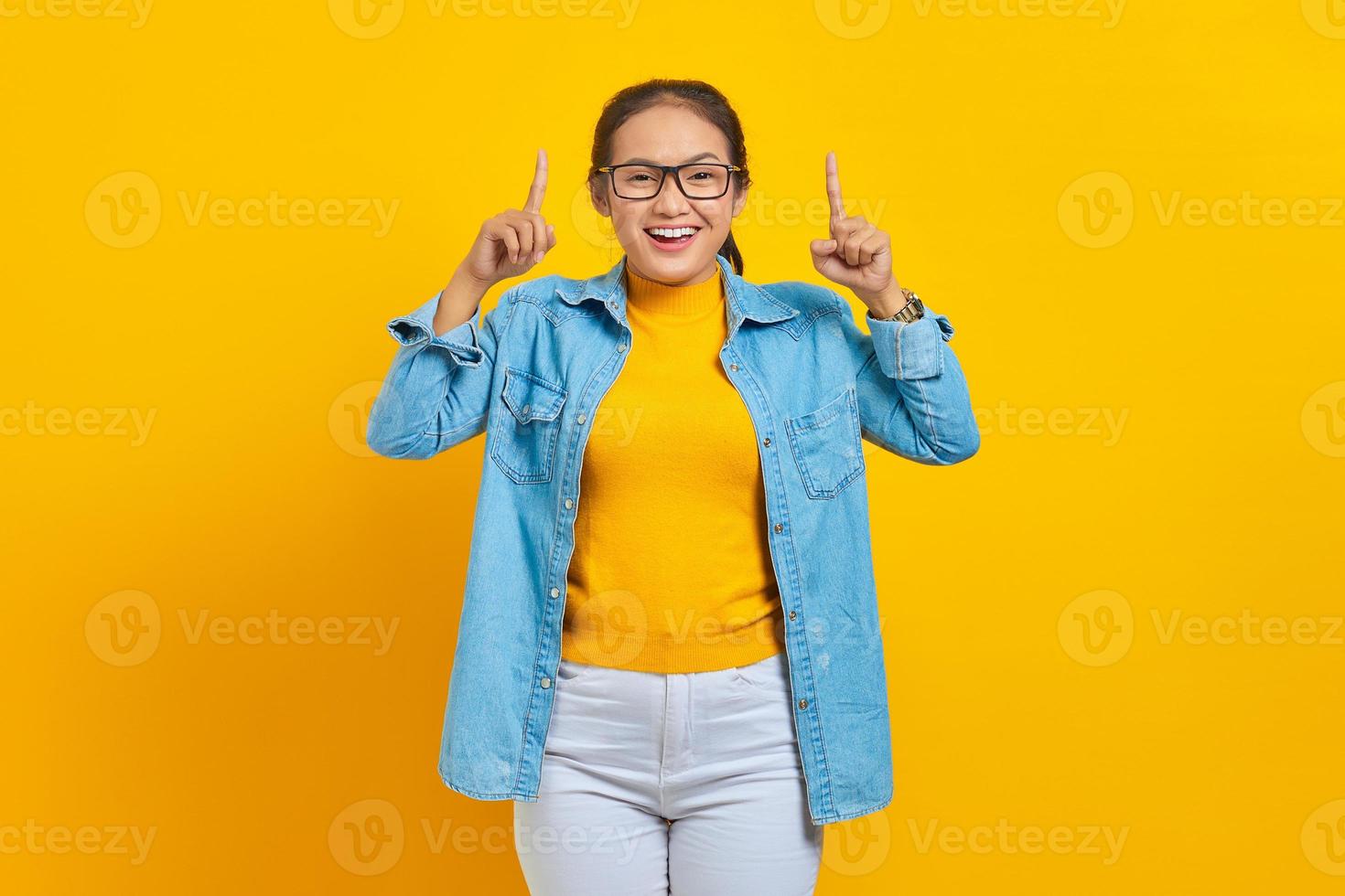 retrato de una joven estudiante asiática sonriente vestida de denim señalando con el dedo hacia arriba, invitando a los clientes a un evento especial aislado en un fondo amarillo. educación en concepto de universidad universitaria foto