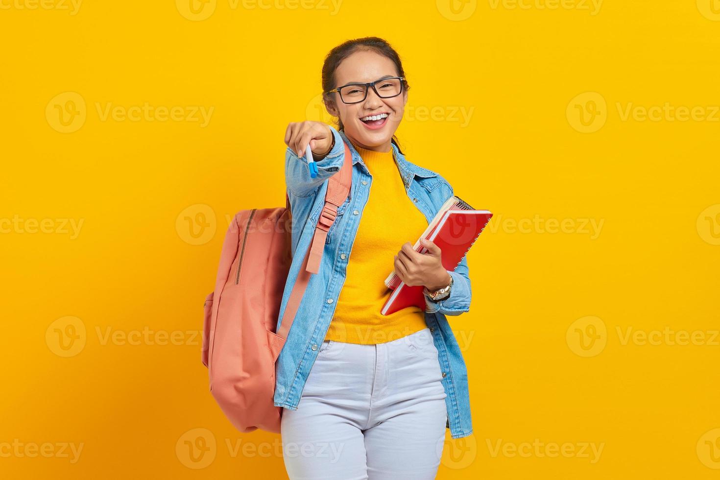 Portrait of cheerful young Asian woman student in casual clothes with backpack holding book and pen, looking at camera isolated on yellow background. Education in college university concept photo