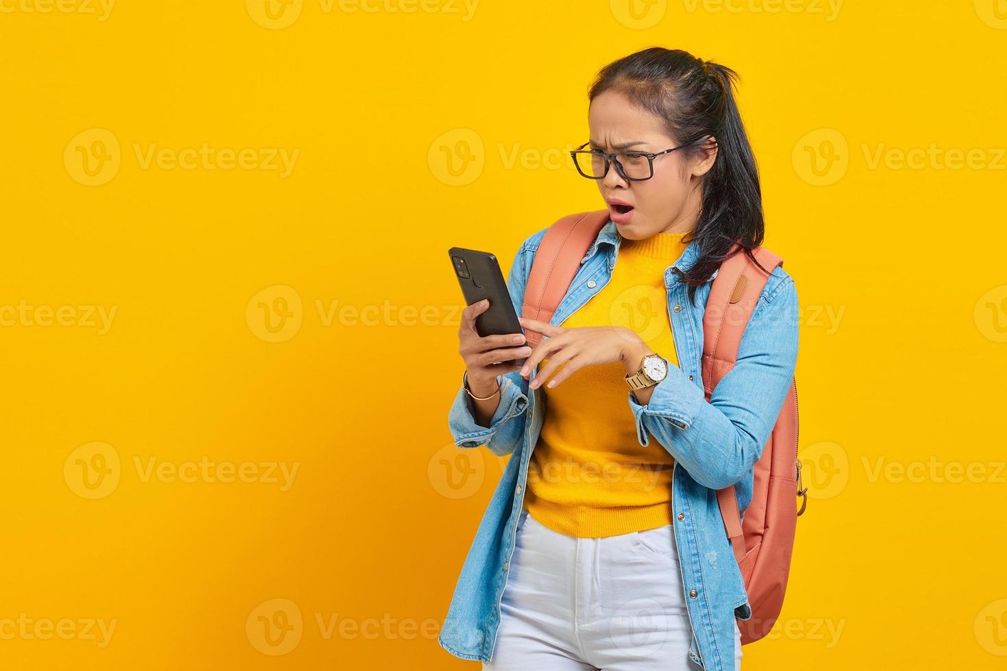 Portrait of  surprised young Asian woman student in casual clothes with backpack typing text message or scrolling feed on social network using smartphone isolated on yellow background photo