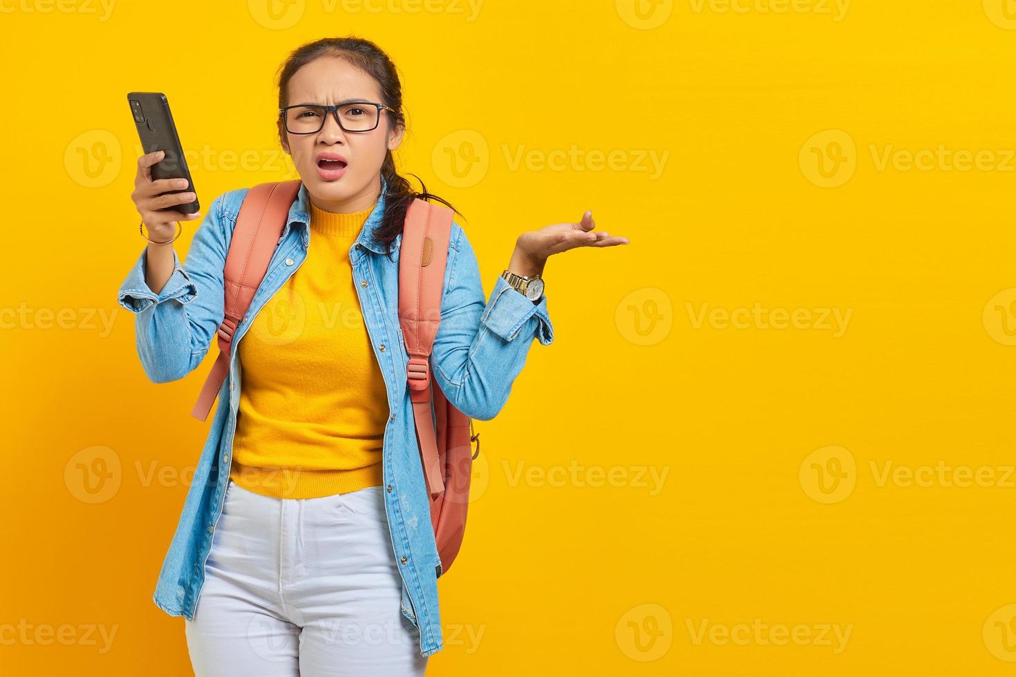 retrato de una joven estudiante asiática confundida con ropa informal con mochila sosteniendo un teléfono móvil y diciendo no con las palmas aisladas en un fondo amarillo. educación en concepto de universidad universitaria foto