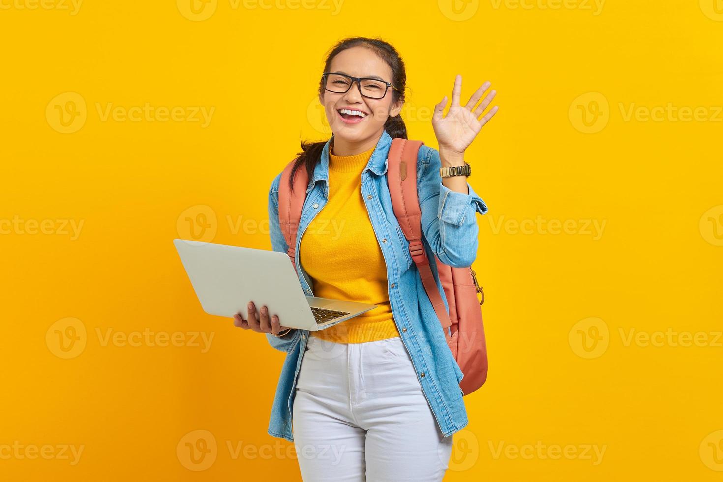 Portrait of cheerful young Asian woman student in casual clothes with backpack using laptop and gesturing waving hand isolated on yellow background. Education in college university concept photo