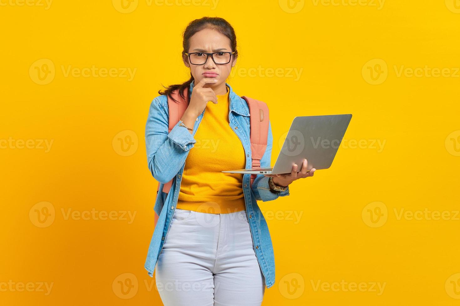 Portrait of serious young Asian woman student in casual clothes with backpack using laptop and thinking about question isolated on yellow background. Education in university college concept photo