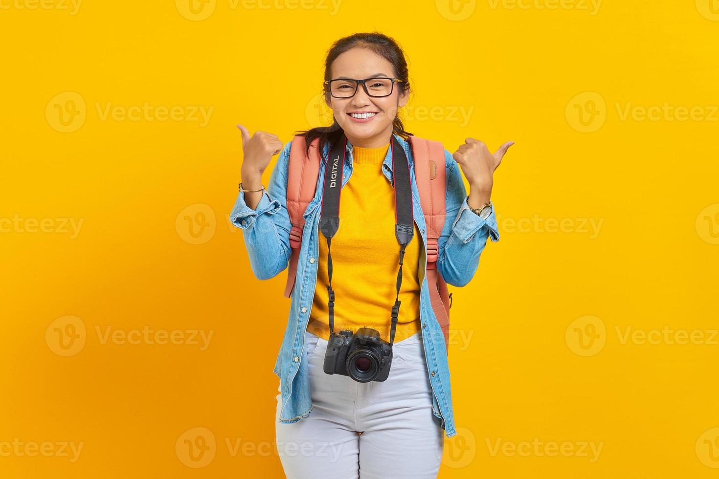 Cheerful young traveler Asian woman with backpack and camera in denim clothes while showing thumb up gesture isolated on yellow background. Passenger traveling on weekends. Air flight journey concept photo