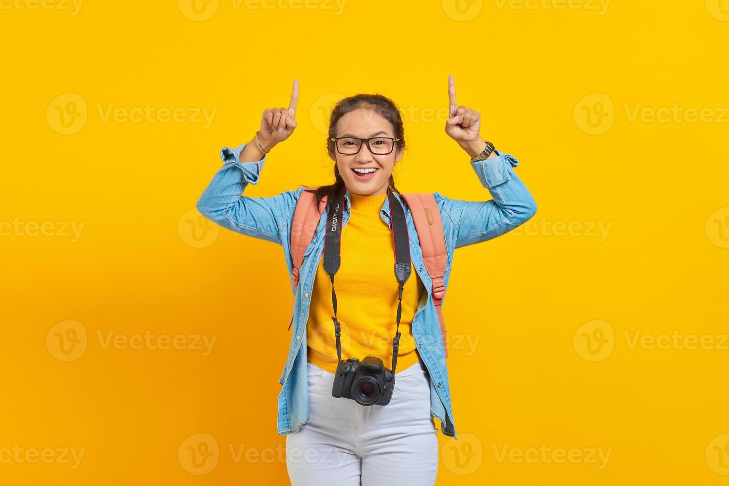 Portrait of cheerful young traveler Asian woman with backpack and camera in denim clothes while pointing at copy space with fingers isolated on yellow background. Air flight journey concept photo