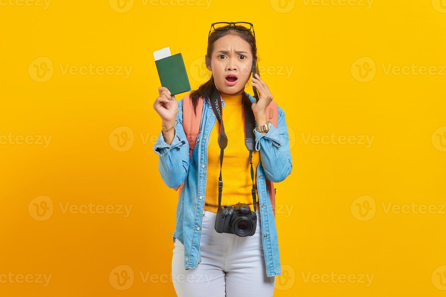 Portrait of surprised young Asian traveler holding boarding pass passport ticket and talking on smartphone isolated on yellow background. Passengers traveling on weekends. Air flight travel concept photo