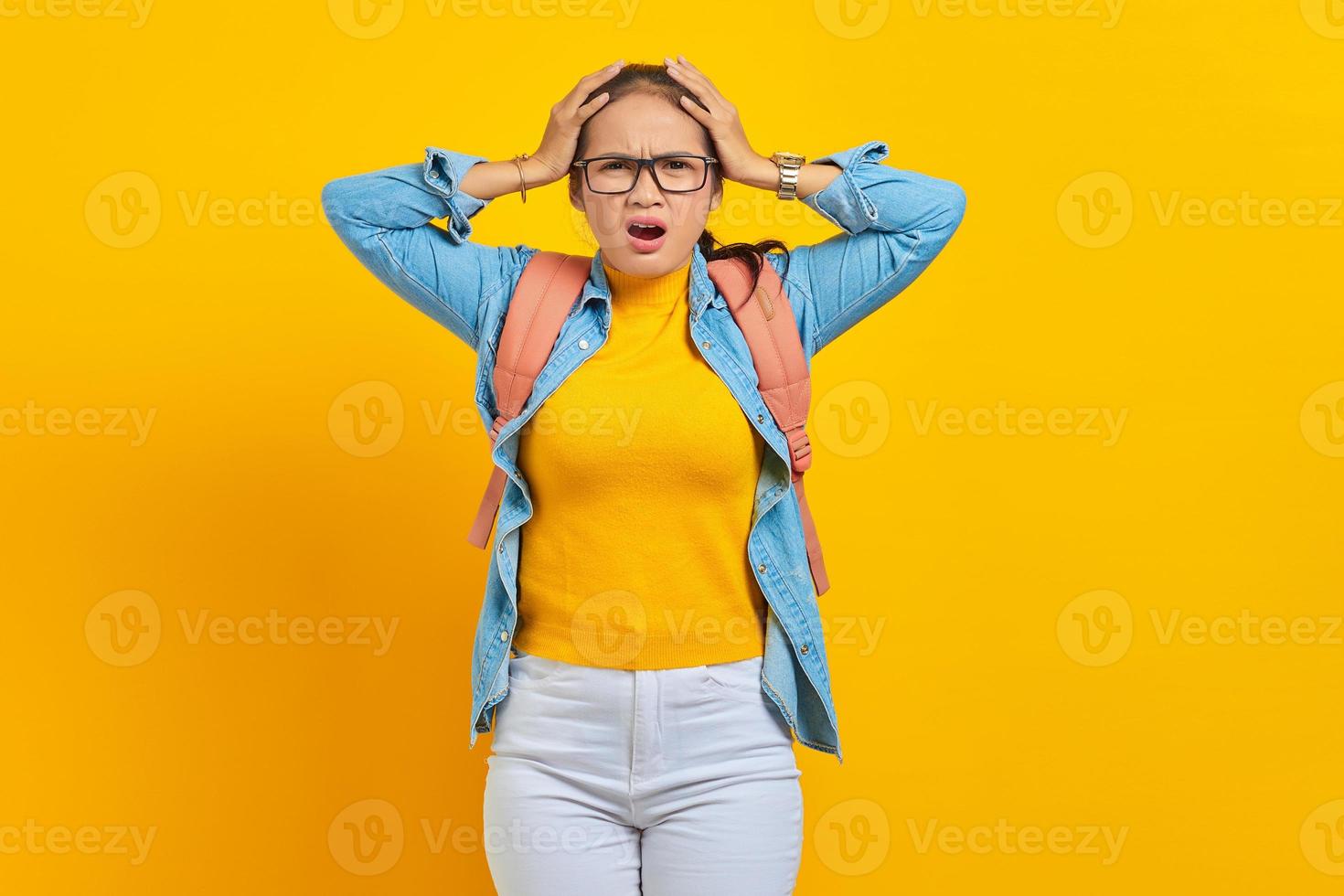 sorprendió a una joven estudiante asiática vestida de denim con mochila, boca abierta y sosteniendo la cabeza en la mano aislada en un fondo amarillo. educación en concepto universitario foto