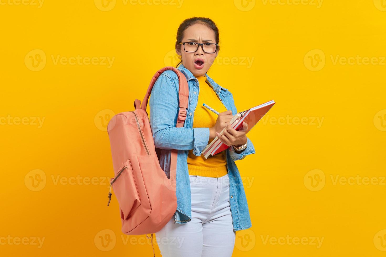 conmocionó a una joven estudiante asiática vestida con ropa de mezclilla con mochila, escribiendo notas en un cuaderno aislado en un fondo amarillo. educación en el concepto de colegio universitario de secundaria foto