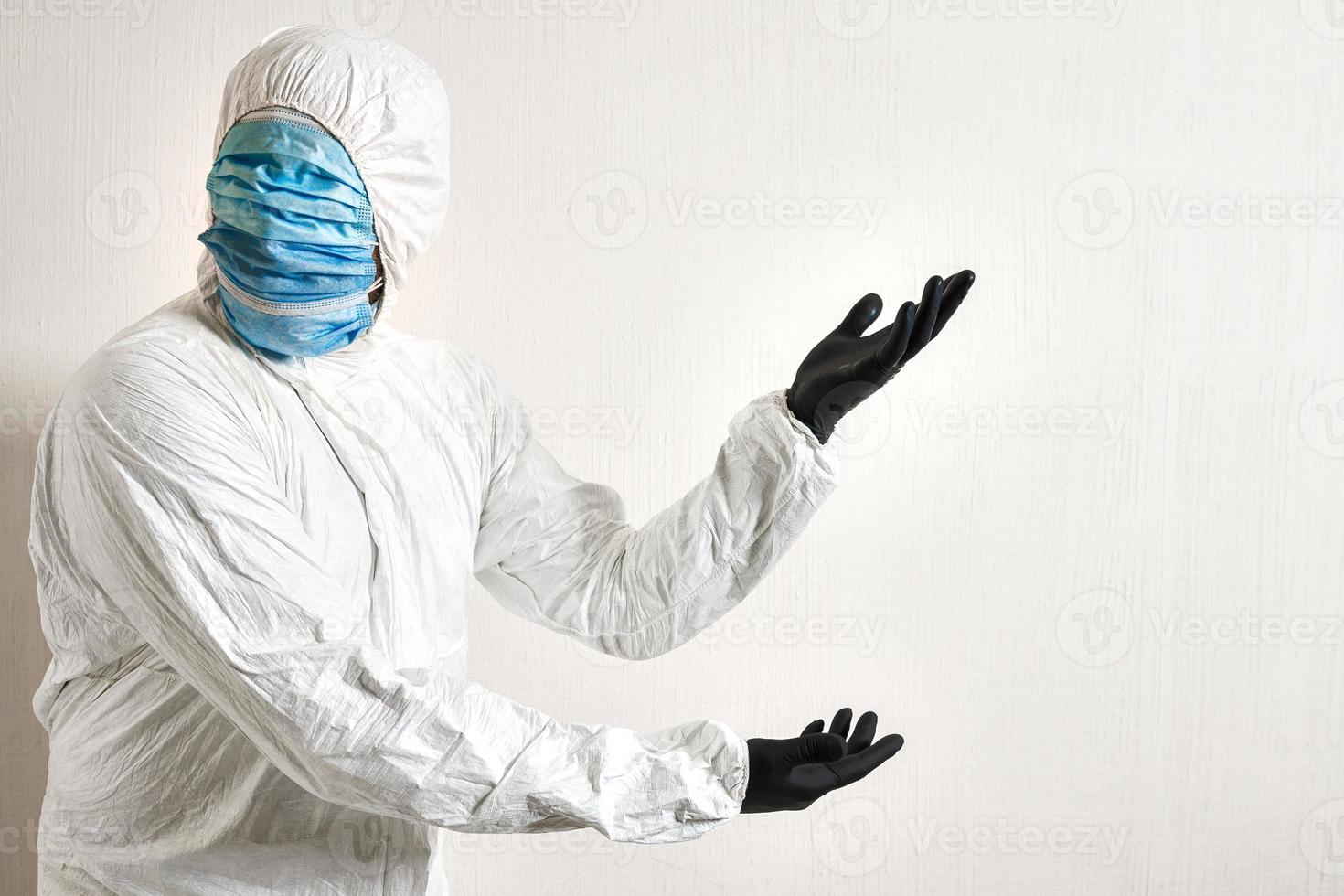 man in a protective suit hung with medical masks posing against wall background showing various gestures with his fingers. the scientist gestures to the wall where the text can be placed photo