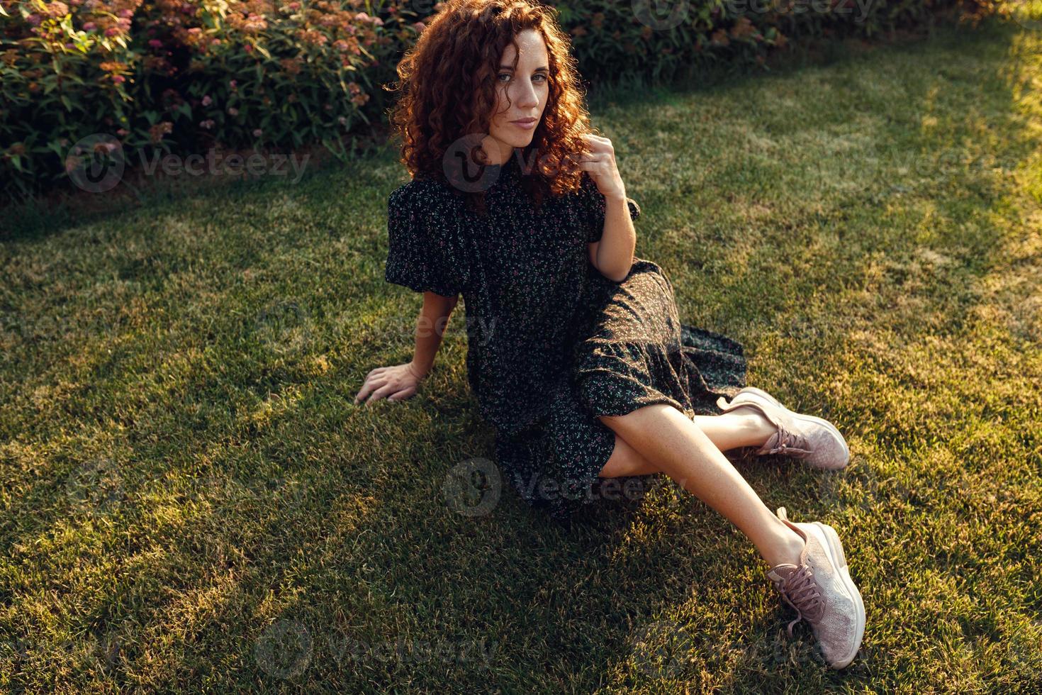 adorable pelirroja rizada con pecas en un vestido disfruta del buen tiempo sentada en el césped foto