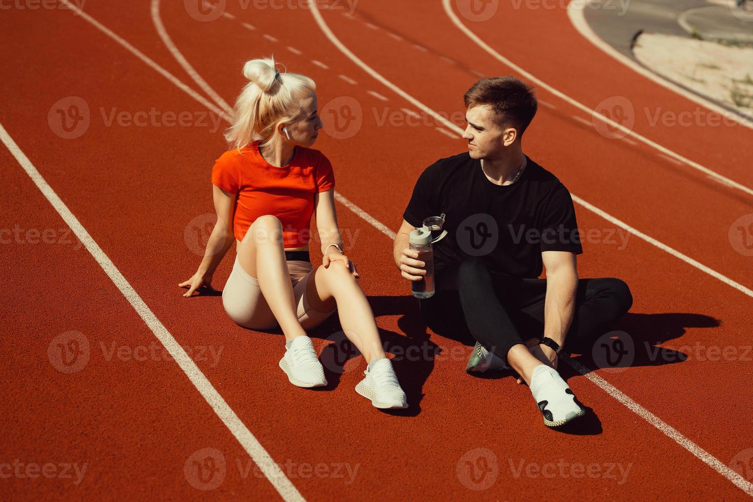 joven hermosa rubia y un chico están sentados en una pista de jogging con una botella de agua y charlando foto