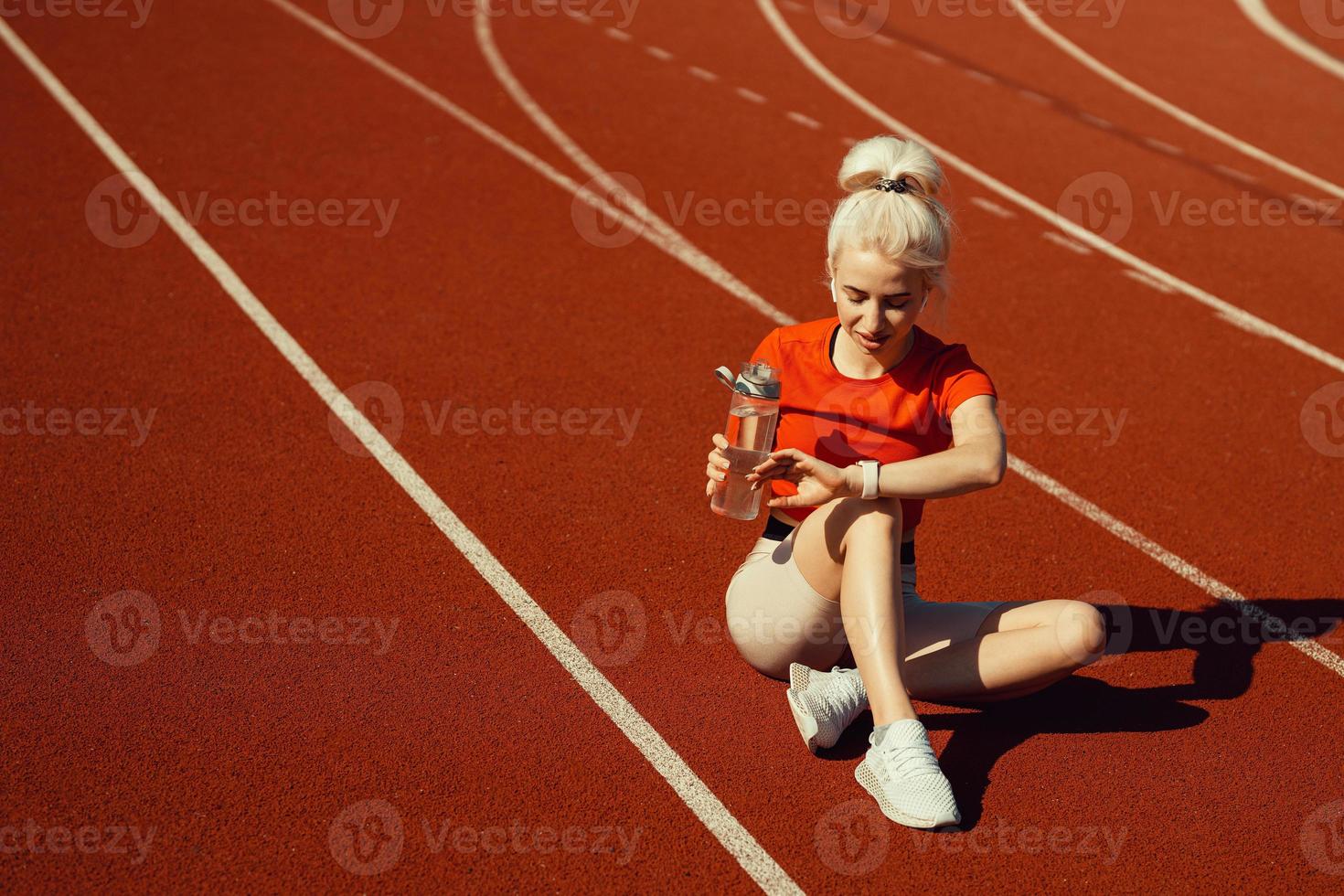 joven y hermosa rubia se sienta en una pista de jogging con una botella de agua y mira su reloj foto