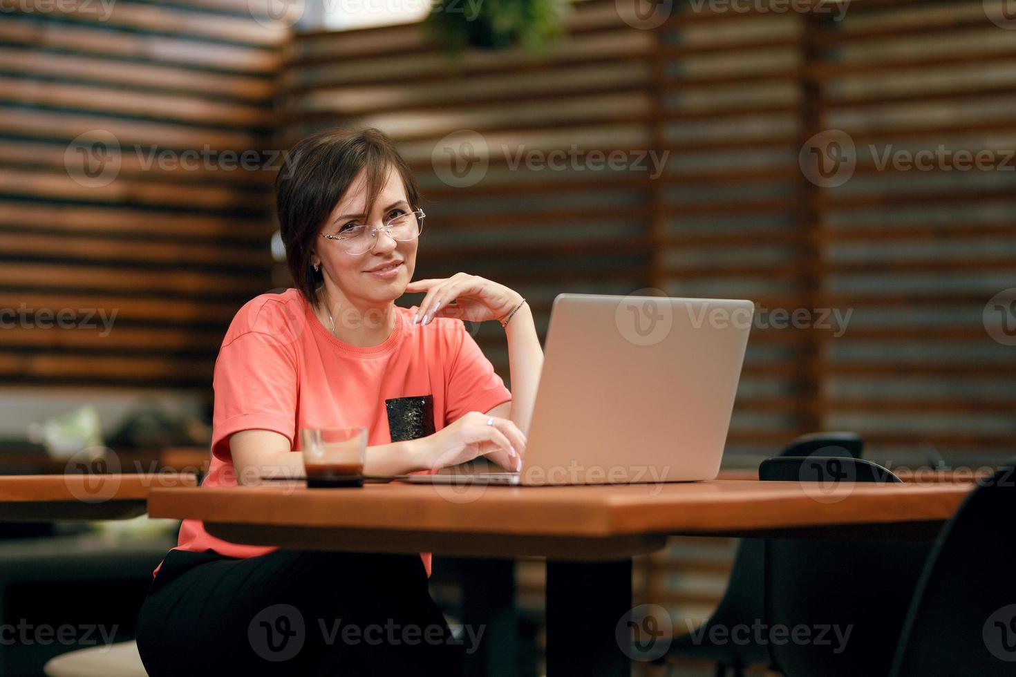 retrato de una mujer profesional madura y segura de sí misma con anteojos, una camiseta de coral sentada en una terraza de verano en un café, usando una computadora portátil para trabajar, riéndose alegremente en el interior foto