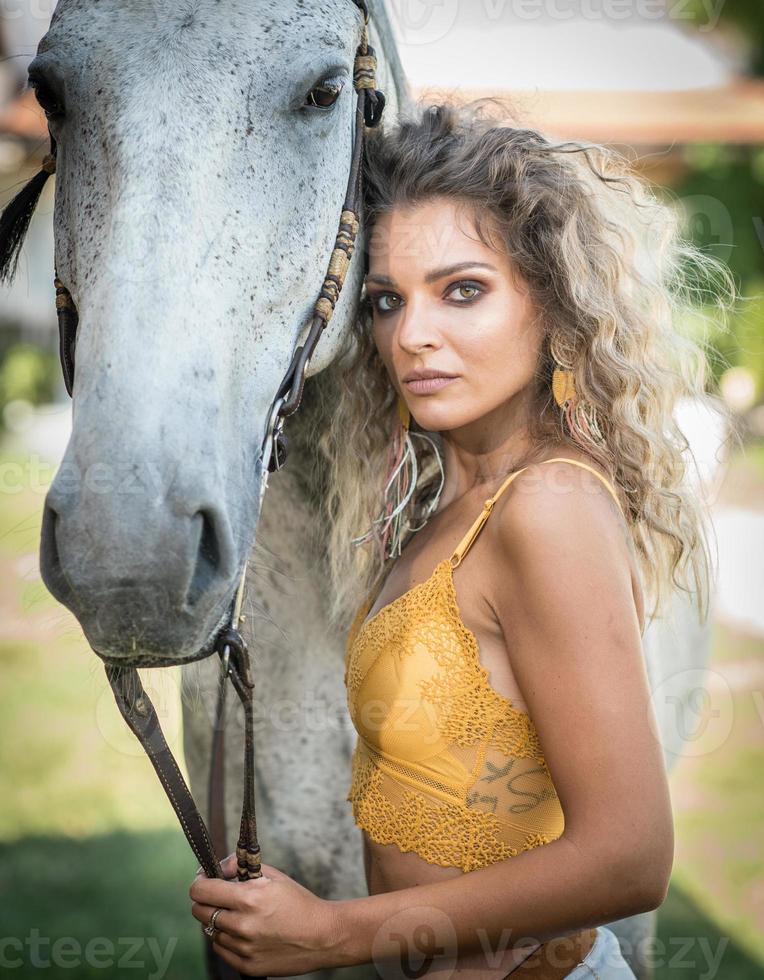 hermosa mujer rubia con cabello rizado y caballo. retrato de una chica con bikini blanco, blusa amarilla transparente y su caballo. bella chica interactuando y divirtiéndose con un caballo en el rancho foto