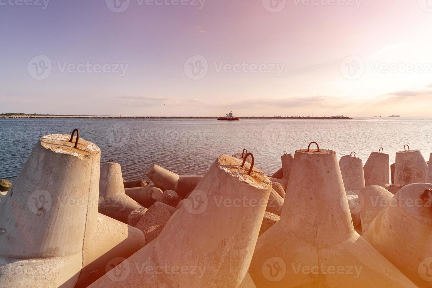Ship-tugboat goes in high seas to tow cargo ship to port, Beautiful sunset over the pier, Tetrapod breakwaters in harbor. photo