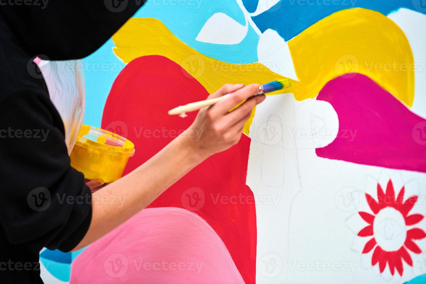artista con capucha negra pintando un cuadro colorido con pincel sobre lienzo blanco en el festival de arte foto