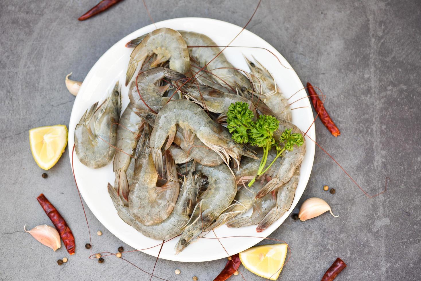 langostinos frescos para cocinar con especias limón sobre fondo oscuro en el restaurante de mariscos, camarones crudos en plato blanco, vista superior foto